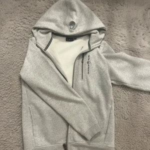 Grå hoodiejacka från Sail Racing - Snygg ljusgrå hoodiejacka från Sail Racing med dragkedja framtill och bröstficka med logga. Jackan har huva, och är tillverkad i mjukt bomullsmaterial. Perfekt för en avslappnad och sportig stil.