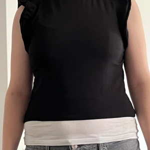 Svart ärmlös topp från 157 - Svart ärmlös topp från 157 med diskreta volangdetaljer vid axlarna. Modellen är figurnära och har rund halsringning. Tillverkad i mjuk bomull med lite stretch för extra komfort. Perfekt att styla med jeans eller kjol. Vita linnet hör ej till!