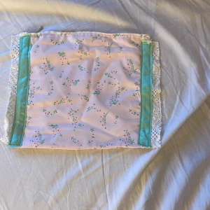 Söt blommig kuvertväska med spets - Pastellrosa kuvertväska i bomull med små gröna och rosa blommor, turkosa band längs sidorna och vit spetskant. Väskan stängs med dragkedja och har en mjuk, romantisk vibe. Perfekt för att bära småsaker eller smink.