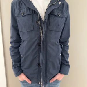 Mörkblå vindjacka från J.Lindeberg - Snygg mörkblå vindjacka från J.Lindeberg med dragkedja och knappar framtill, två bröstfickor med lock och knapp samt emblem på ärmen. Jackan har en clean look och är tillverkad i lätt polyester, perfekt för vår och höst.