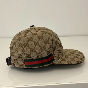 Gucci keps med GG-mönster och ränder - Snygg keps från Gucci i beige och brunt med klassiskt GG-monogram över hela kepsen. På sidan finns den ikoniska gröna och röda randen. Justerbar rem baktill och insida i brunt tyg. Perfekt för dig som vill sticka ut med lyxig streetstyle.