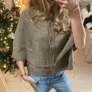 Beige stickad poncho med knappar - Säljer en beige stickad kofta med stora knappar framtill. Koftan har korta, vida ärmar och ribbade kanter vid ärmslut och nederkant. 