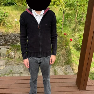 Svart stickad cardigan jacka Tommy Hilfiger - Svart stickad cardigan jacka från Tommy Hilfiger med dragkedja framtill och ribbade muddar. Liten logga på bröstet och snygg krage med vinröd insida. Perfekt för lager-på-lager och chill höstkvällar.