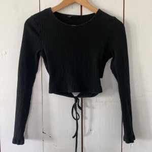 Svart ribbad croppad topp med knyt - Svart långärmad croppad topp med ribbad struktur och knytband i ryggen. Toppen har rund halsringning och är figurnära i modellen. Perfekt för att skapa en snygg och trendig look till jeans eller kjol.
