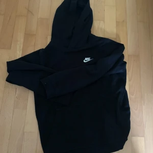 Svart Nike hoodie med logga - Svart hoodie från Nike med klassisk vit broderad logga på bröstet. Tröjan har huva och en stor magficka framtill. Perfekt för en chill och sportig stil. Mjuk och skön bomullsblandning, långärmad och med relaxed fit.