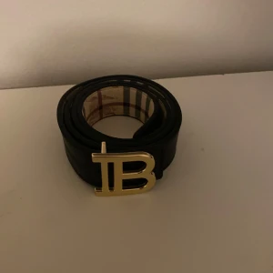 Burberry bälte - Snyggt svart skärp från Burberry med klassiskt rutigt mönster på insidan och präglat mönster på utsidan. Stort guldigt 'B'-spänne framtill som ger en lyxig känsla. Perfekt accessoar för att lyfta din outfit. Fraktar inom 24h!