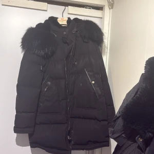 Svart dunjacka med fuskpäls från Parajumpers - Snygg svart dunjacka från Parajumpers med stor fluffig fuskpäls runt huvan. Jackan har dragkedja, stora fickor med läderdetaljer och en cool, lite oversized look. Perfekt för kalla dagar när du vill vara både varm och stilren. Pris kan diskuteras 