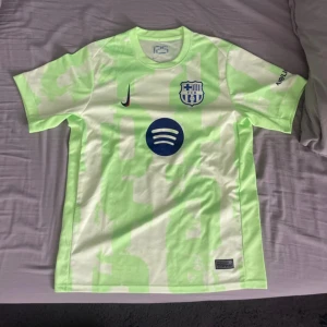 FC Barcelona Nike fotbollströja L - Säljer en FC Barcelona fotbollströja från Nike i ljusgrön och vit camo-design. Tröjan har klubbmärke, Nike-logga och Spotify som sponsor framtill samt UNHCR-tryck på ryggen. Tillverkad i Dri-FIT-material som andas, perfekt för träning eller match. Använd endast en gång