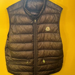 Brun dunväst från Moncler - Snygg brun dunväst från Moncler med klassisk quiltad design och dragkedja framtill. Västen har två fickor med dragkedja och ikonisk Moncler-logga på bröstet. Perfekt för lager-på-lager och ger en clean look till din outfit.