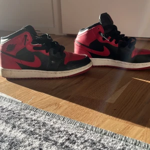 Nike Air Jordan 1 Mid Svart/Röd 36.5 - Säljer ett par ikoniska Nike Air Jordan 1 Mid i svart och rött med klassisk design och snygga detaljer. Skorna har svart snörning, röd swoosh och perforerad tå. Tillverkade i skinn och textil, perfekta för dig som gillar streetstyle.