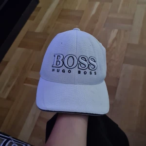 Vit keps från Hugo Boss - Snygg vit keps från Hugo Boss med stor svart broderad logga framtill. Kepsen har justerbart spänne bak med metallplatta och svart randig detalj. Tillverkad i luftig bomull med klassisk böjd skärm. Perfekt för en clean och sportig look.