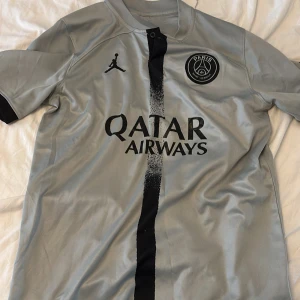 PSG x Jordan grå fotbollströja - Snygg grå PSG fotbollströja från samarbetet med Jordan. Tröjan har svart tryck med Qatar Airways-logga, PSG-emblem på bröstet och en svart vertikal rand i mitten. Tillverkad i lätt och ventilerande material, perfekt för träning eller match.