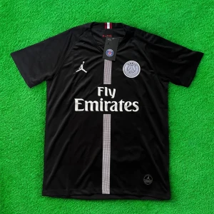 Svart PSG fotbollströja Jordan x Fly Emirates - Svart PSG fotbollströja med korta ärmar, tillverkad i lätt och ventilerande material. Framsidan har vit vertikal rand, broderad PSG-logga och Jumpman-logga samt Fly Emirates-tryck. Rödvit detalj i nacken. Perfekt för dig som älskar fotboll och streetstyle.