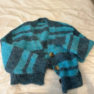 Randig blå kofta från Gina Tricot - Mysig stickad kofta från Gina Tricot i blå och turkosa ränder. Koftan har stora träknappar framtill, lång ärm och en loose fit. Perfekt för lager-på-lager och chill dagar. Materialet är fluffigt och ger en cool vintagekänsla.