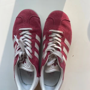 Adidas Gazelle rosa mockasneakers - Adidas Gazelle sneakers i rosa mocka med vita detaljer. Skorna har vit platt sula, rund tå och snörning. 