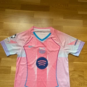 FC Barcelona rosa matchtröja Nike - Säljer en FC Barcelona fotbollströja i rosa och lila toner från Nike. Tröjan har korta ärmar, V-ringning med blå detalj, vita linjemönster och tryckta loggor från Spotify och UNHCR. Materialet är lätt och andas, perfekt för fotboll eller att bära som supporter.