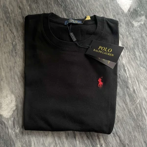 Svart t-shirt från Polo Ralph Lauren - Svart t-shirt från Polo Ralph Lauren med klassisk rund hals och korta ärmar. Ikoniska röda broderade loggan på bröstet ger en clean och stilren look. Tillverkad i mjuk bomull som känns skön mot huden. Perfekt för dig som gillar enkel men snygg stil.