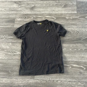 Svart t-shirt från Lyle & Scott - Svart t-shirt från Lyle & Scott med klassisk rund hals och korta ärmar. På bröstet finns den ikoniska gula örnloggan broderad. Skön och enkel modell i mjuk bomull, perfekt till jeans eller shorts.