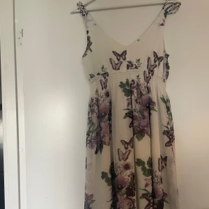 Ljus blommig långklänning med fjärilar - Romantisk långklänning i ljus ton med lila blommor och fjärilar. Klänningen har smala axelband, markerad midja med knytband bak och ett luftigt, svepande fall. Perfekt för sommarens alla tillfällen. Materialet känns lätt och svalt. Använt några gånger så skicket är väldigt bra. Står ingen storlek men skulle säga att det är som en xs. Går att diskutera pris 