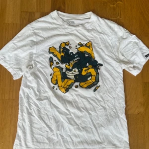 Vit Vans t-shirt - Vit t-shirt från Vans med ett stort grafiskt tryck på bröstet i svart och gult. Klassisk passform och rund halsringning. Tillverkad i 100% bomull, perfekt för dig som gillar streetwear och coola prints.