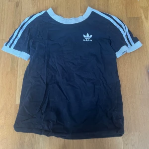 Svart Adidas t-shirt med blå ränder - Svart t-shirt från Adidas med vita ribbade muddar och klassiska tre ränder längs ärmarna. Adidas-logga på bröstet. Skön passform och sportig stil, perfekt för dig som gillar streetwear. Materialet är mjuk bomull.