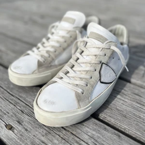 Philippe Models - Snygga vita sneakers från Philippe Model med detaljer i ljusgrå mocka och klassisk snörning. Skorna har en rund tå, platt sula och den ikonisk sköldloggan på sidan. De är mycket sparsamt använda och är i toppskick det finns en liten defekt på insidan av skon men det är inget som syns när man har på sig skon. Yttermaterialet är en mix av skinn och mocka för en stilren och trendig look. Perfekt inför skolstart! Det ingår en Phillipe model dustbag. Pris går att diskutera.