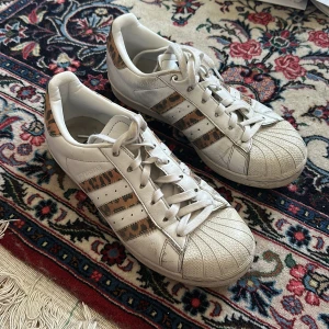 Adidas Superstar med leoparddetaljer - Snygga vita Adidas Superstar sneakers med klassisk snörning och coola leopardmönstrade detaljer på de ikoniska tre ränderna och hälen. Skorna har rund tå, platt sula och är tillverkade i skinn. Perfekta för dig som vill sticka ut med en unik twist på en tidlös modell.