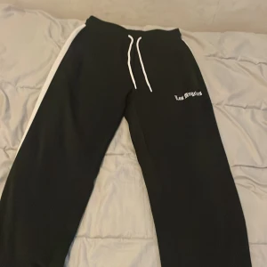 Svarta mjukisbyxor Los Angeles - Svarta mjukisbyxor med vit text 'Los Angeles' på ena benet och vita snören i midjan. Snygg vit rand längs sidan ger en sportig vibe. Perfekta för chill eller streetwear. Mjukt material och relaxed fit.