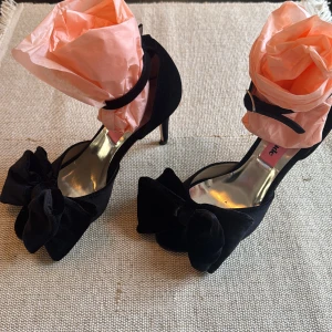 Svarta pumps med rosett från Custommade - Stilrena svarta pumps i sammet från Custommade med hög klack och öppen tå. Skorna har en stor dekorativ rosett framtill och smal ankelrem med spänne. Perfekta för dig som vill sticka ut med en elegant och trendig look.