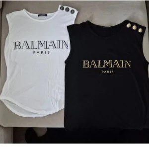 Balmain Paris ärmlösa toppar med knappar - Två ärmlösa toppar från Balmain Paris, en i vitt med svarta tryck och silvriga lejonknappar på axeln, och en i svart med guldigt tryck och guldknappar. Båda har rund halsringning och är i mjuk bomull. Perfekta för en trendig och lyxig look. ÄKTA
