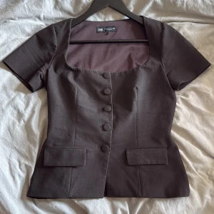 Brun kortärmad topp från Zara - Snygg brun topp från Zara med korta ärmar och markerad midja. Toppen har en fyrkantig urringning, klädda knappar framtill och två dekorativa ficklock. Perfekt för dig som gillar en stilren och chic look.