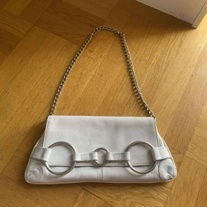 Vit axelväska i skinn från Zoul - Snygg vit axelväska från Zoul i skinn med coola silverfärgade ringdetaljer på framsidan och en kedjerem. Väskan har ett innerfack med dragkedja. 🩷