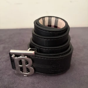 Svart Burberry bälte med silverspänne - Stilrent svart bälte från Burberry med klassiskt rutmönster och stort silverspänne med 'B'-logga. Bältet är gjort i skinn och har en snygg detaljrik finish med diskret Burberry-emblem. Insidan har den ikoniska Burberry-rutan i beige, svart och rött.