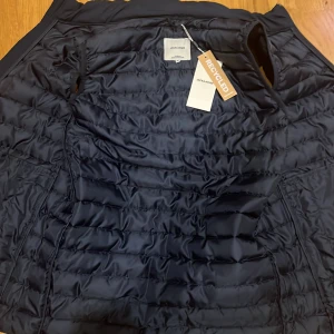 Marinblå dunväst från Jack & Jones - Marinblå  dunväst från Jack & Jones med quiltad design och dragkedja framtill. Västen är tillverkad i återvunnet syntetmaterial och har en stilren, enkel look med hög krage. Perfekt att slänga över en hoodie eller tröja för extra värme.