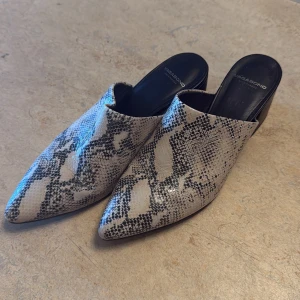 Ormskinnsmönstrade Vagabond mules - Snygga mules från Vagabond med spetsig tå och låg klack. Skorna har ett ormskinnsmönster i beige och svart, vilket ger en edgy vibe. Tillverkade i skinn med svart innersula och öppen häl. Perfekta för dig som vill sticka ut.