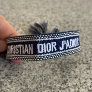 Blått Christian Dior J'Adior armband - Snyggt blått armband från Christian Dior med texten 'J'Adior' broderad i vitt. Armbandet har justerbar knytning med två tofsar i ändarna och är gjort i ett mjukt tygmaterial. Perfekt accessoar för att lyfta din outfit.