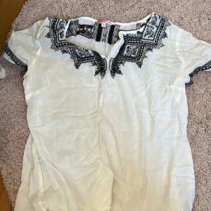 Vit blus med svart broderi Indiska - Säljer en vit blus från Indiska i storlek M med snygga svarta broderade detaljer runt halsringningen och på ärmarna. Blusen har korta ärmar och en avslappnad passform, perfekt för en chill och bohemisk stil.