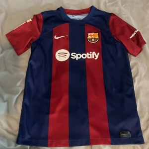 FC Barcelona Lewandowski tröja Nike - Säljer en officiell FC Barcelona matchtröja från säsongen 2022/2023 med Lewandowski och nummer 9 på ryggen. Tröjan är blå och röd med breda ränder, korta ärmar och tillverkad i Dri-FIT material. Har klubbmärke, Nike-logga, Spotify sponsor och La Liga-märke på ärmen.