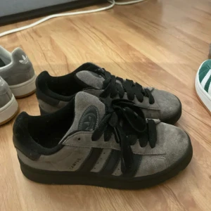 Gråa Adidas Campus sneakers i mocka - Säljer ett par gråa Adidas Campus sneakers med svarta detaljer och klassiska tre ränder på sidan. Skorna har svart sula, svart snörning och är tillverkade i mjuk mocka. Perfekta för dig som gillar en clean och stilren look.