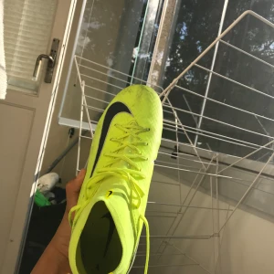 Nike Mercurial gula fotbollsskor - Säljer ett par Nike Mercurial fotbollsskor i neon-gult med svarta detaljer och silvergrå häl. Skorna har snörning, högre strumpliknande skaft och är tillverkade i syntetmaterial för bästa passform och grepp på planen. Perfekta för dig som vill sticka ut på matchen.