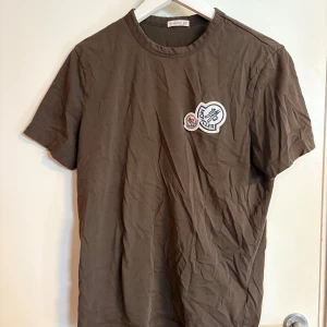 Brun-grön Moncler t-shirt med patch - Snygg brun t-shirt från Moncler med klassisk rund hals och korta ärmar. På bröstet sitter två broderade Moncler-patchar som ger en exklusiv vibe. Tillverkad i mjuk bomull som känns skön mot huden. Perfekt för dig som gillar stilrena plagg med en twist. Skick-8/10 då lappen är någorlunda sliten. Annars felfri. Priset är självklart förhandlingsbart 