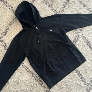 Svart hoodie från Polo Ralph Lauren - Svart hoodie från Polo Ralph Lauren med klassisk vit broderad logga på bröstet. Tröjan har dragkedja framtill, huva med snörning och två stora fickor. Tillverkad i mjuk bomull, perfekt för en chill och stilren look.