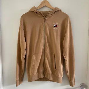 Beige hoodie från Tommy Hilfiger - Snygg beige hoodie från Tommy Hilfiger med dragkedja, huva och klassisk logga broderad på bröstet. Tröjan har ribbade muddar, kängurufickor och en cool randig detalj på huvan. Perfekt för en chill och stilren look.
