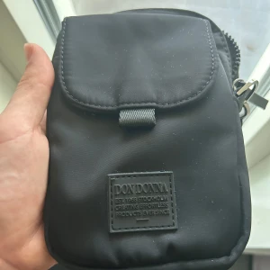 Svart axelväska från Don Donna - Säljer en stilren svart axelväska från Don Donna med justerbar rem och praktiskt lock med kardborre. Väskan har dragkedja upptill och en diskret logotyp framtill. Perfekt storlek för mobil, plånbok och andra småsaker. Enkel och clean design.
