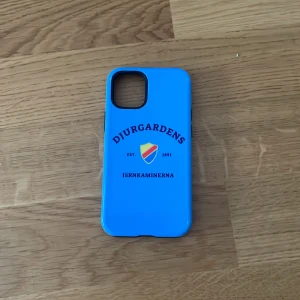 Blå Djurgården mobilskal i plast - Snyggt blått mobilskal till iPhone 12 med Djurgårdens IF-logga och texten Järnkaminerna på baksidan. Skalet är gjort i tålig plast och har öppningar för kamera och knappar. Perfekt för dig som vill visa vilket lag du håller på!