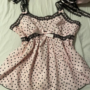 Rosa hjärtmönstrad linnetopp med spets - Supersöt rosa babydoll topp med svarta hjärtan, dekorerad med svart spets och en söt rosett framtill. Axelbanden är knytbara med spetsdetaljer och toppen har en flowy passform