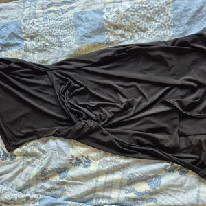 Svart långklänning med drapering - Snygg svart långklänning från Nelly med en axel och draperad detalj framtill. Klänningen har en elegant, figurnära passform och är tillverkad i ett mjukt, stretchigt material som ger en lyxig känsla. Perfekt för dig som vill sticka ut med en stilren look.