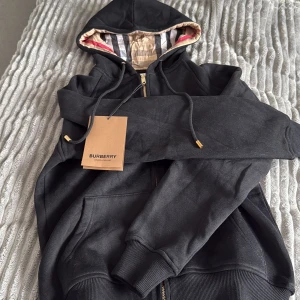 Svart Burberry hoodie med dragkedja -  Helt ny svart hoodie från Burberry med klassiskt rutigt foder i huvan och guldfärgade detaljer på snörningen. Tröjan har dragkedja framtill, fickor och ribbade muddar. Perfekt för dig som vill ha en stilren och lyxig streetwear-look.