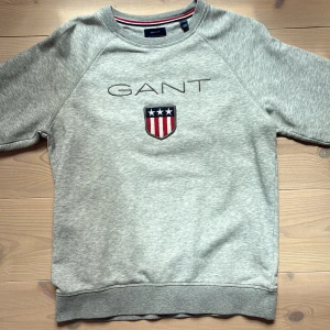 Grå sweatshirt från GANT med tryck - Snygg grå sweatshirt från GANT med broderad logga och sköld på bröstet. Tröjan har rund hals, långa ärmar och ribbade muddar. Perfekt för en chill och sportig look. Tillverkad i mjuk bomullsmix för extra komfort. Pris kan diskuteras vid snabb affär. 