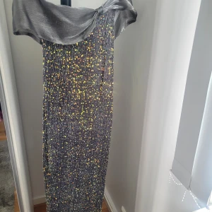 Glittrig långklänning med offshoulder - Snygg långklänning i silver med glittriga paljetter i flera färger. Klänningen har en bred offshoulder-krage och är figurnära. Perfekt för dig som vill sticka ut på festen!